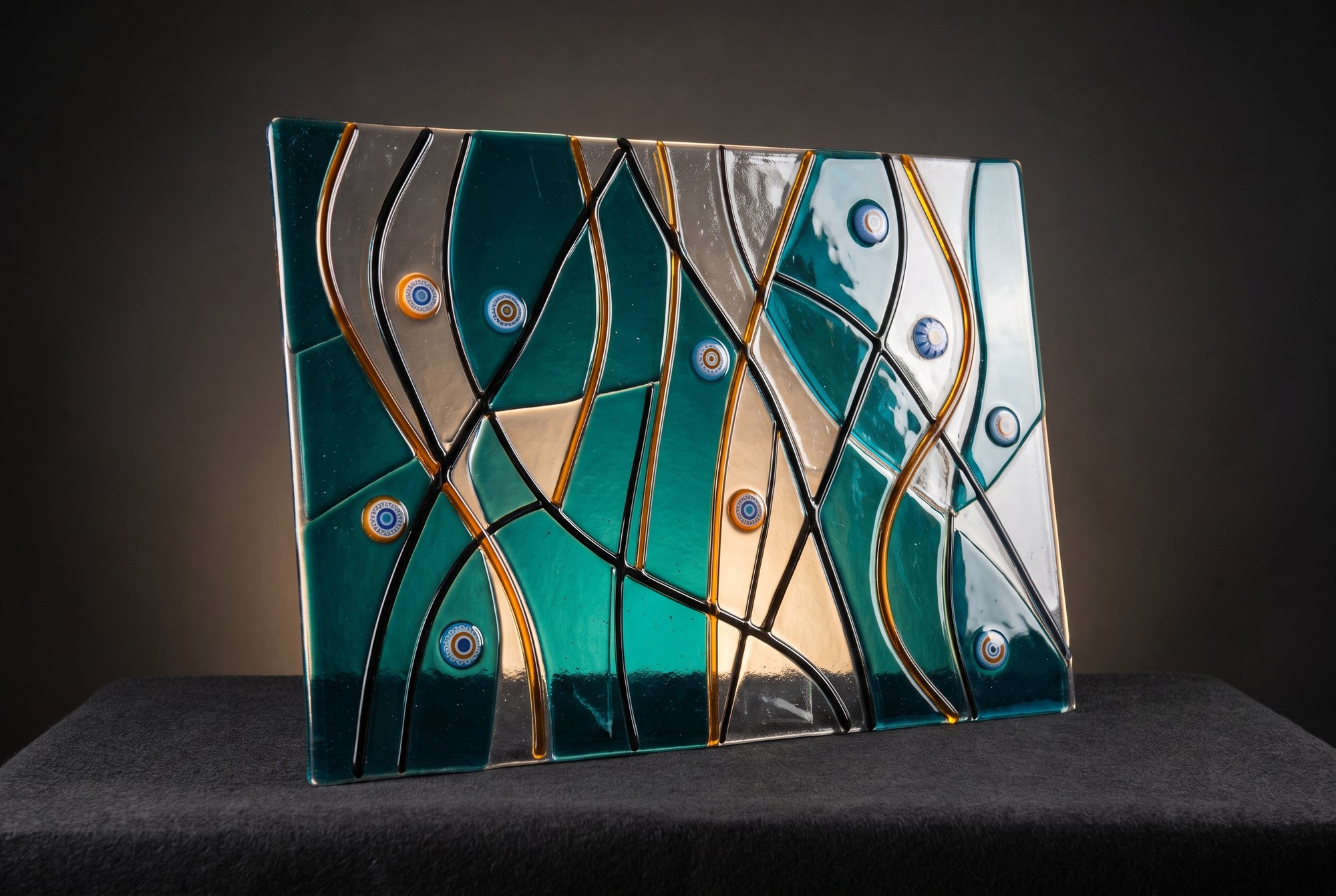 A fused glass panel of teal and amber ribbons with inlaid murrine discs, lit from behind